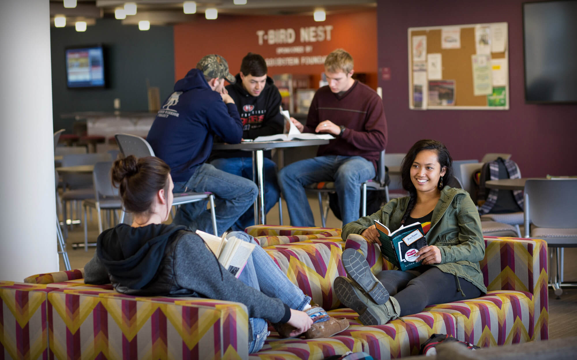 casper-college-04-t-bird-nest Students sitting on couches with books and at high-top tables studying with brightly painted walls in background.