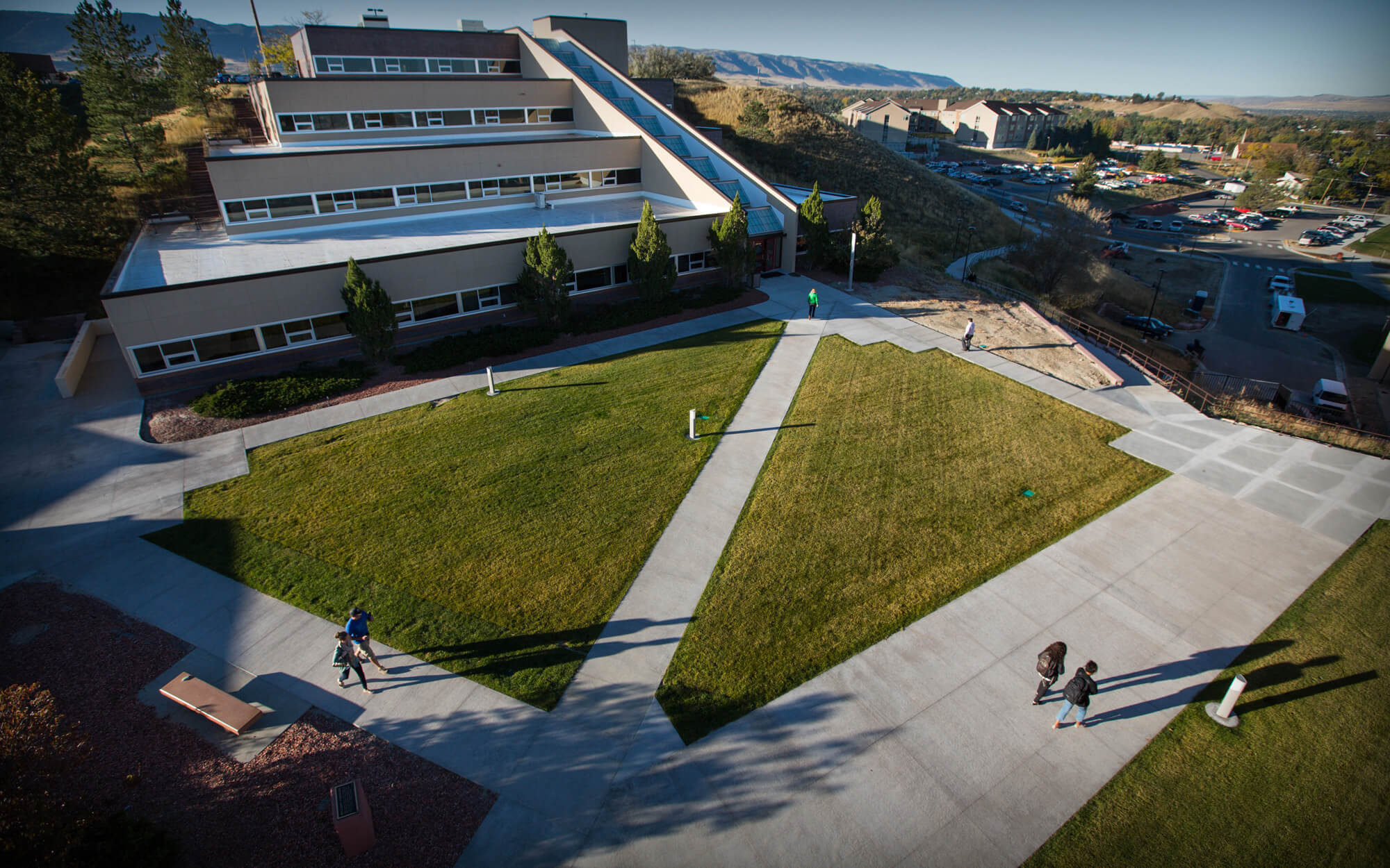 Aerial photo of students walking along sidewalk next to green grass spaces in front of a four-level building.