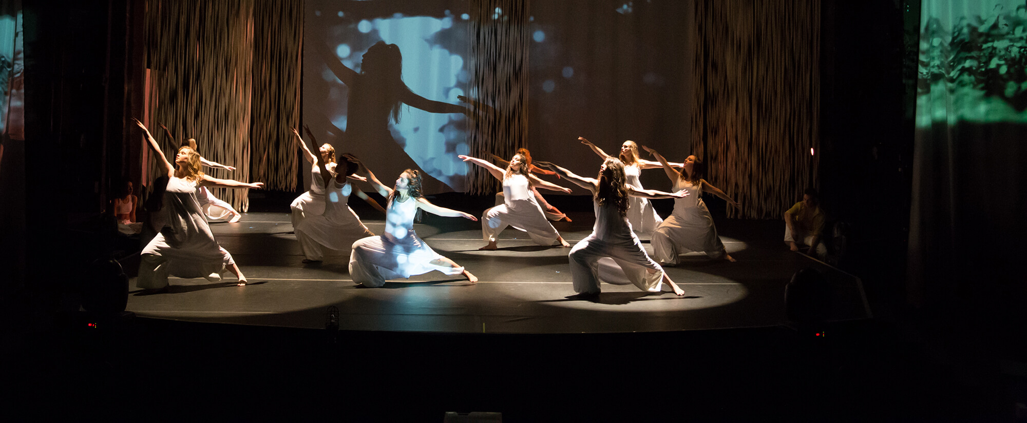 A group of dancers dressed in loose white clothing on a dimly lit stage with a spotlight on them.