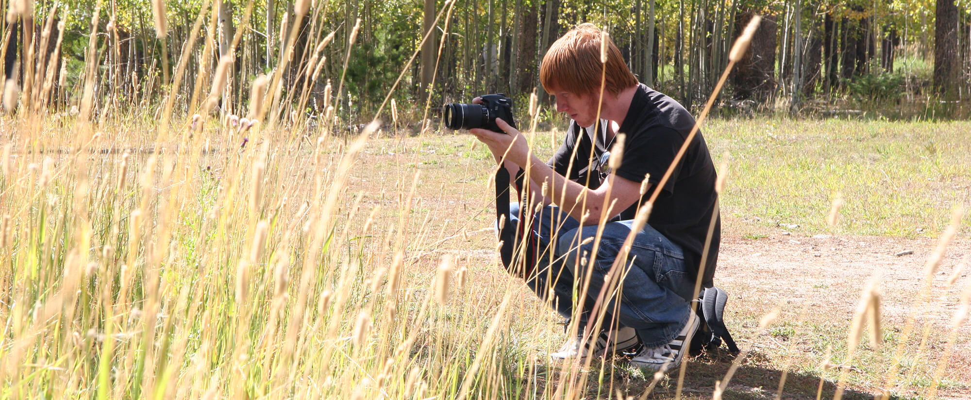 Student holding a DSLR camera crouched in tall grass on top of a mountain.