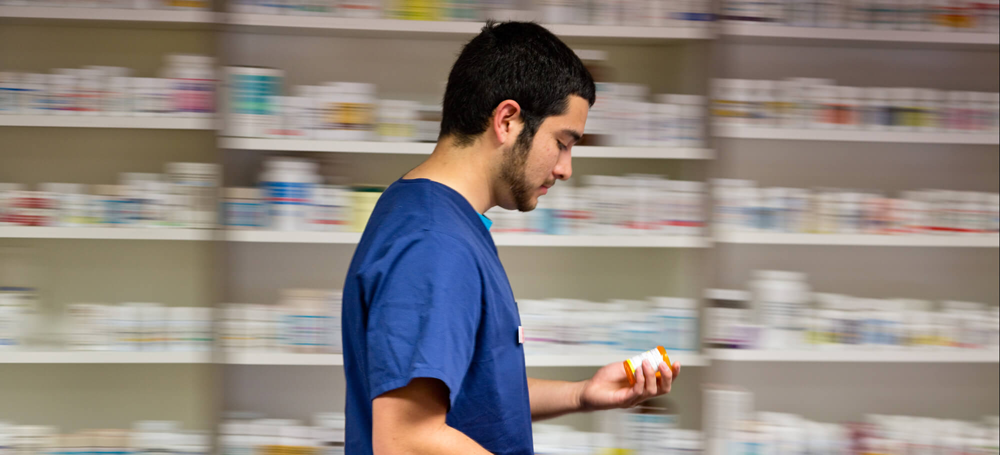 A student holds a pill bottle while walking past shelves set up like a pharmacy.