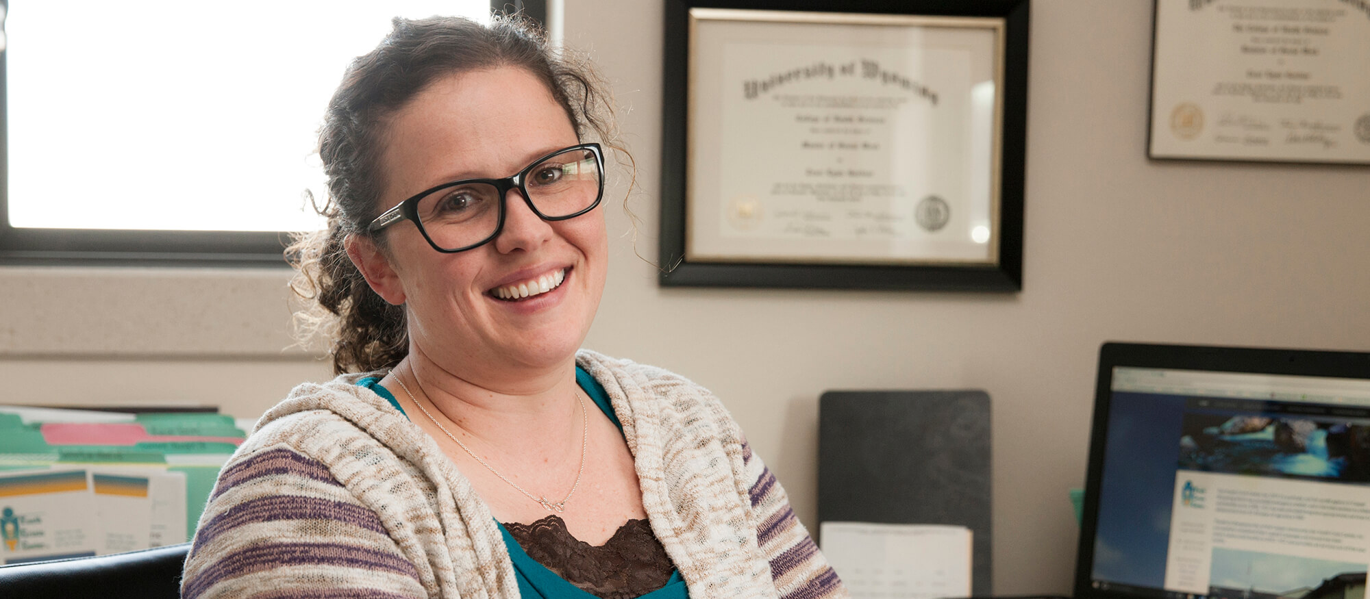 accounting-casper-college Casper College grad Traci Blevins sits at her work desk with a diploma on the wall behind her and a file rack of folders behind her.