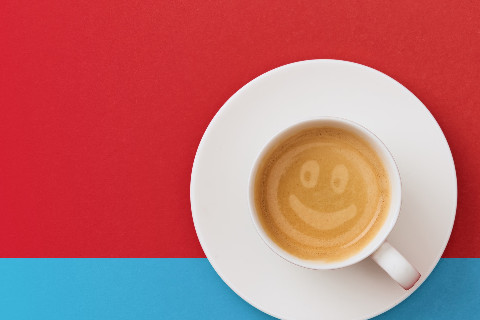 Photo of a white coffee cup that holds a coffee drink with a smiley face in the foam on a red and turquoise background