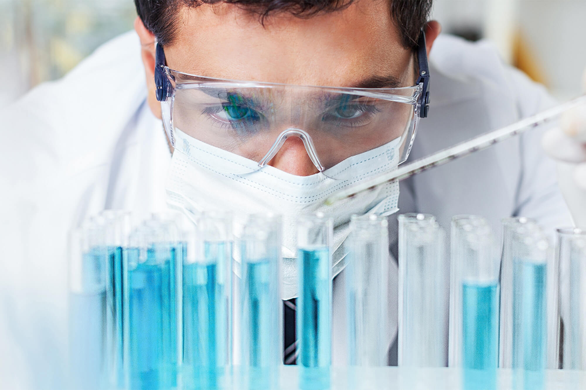 Photo of man with safety glasses and face mask with test tubes filled with blue liquid.