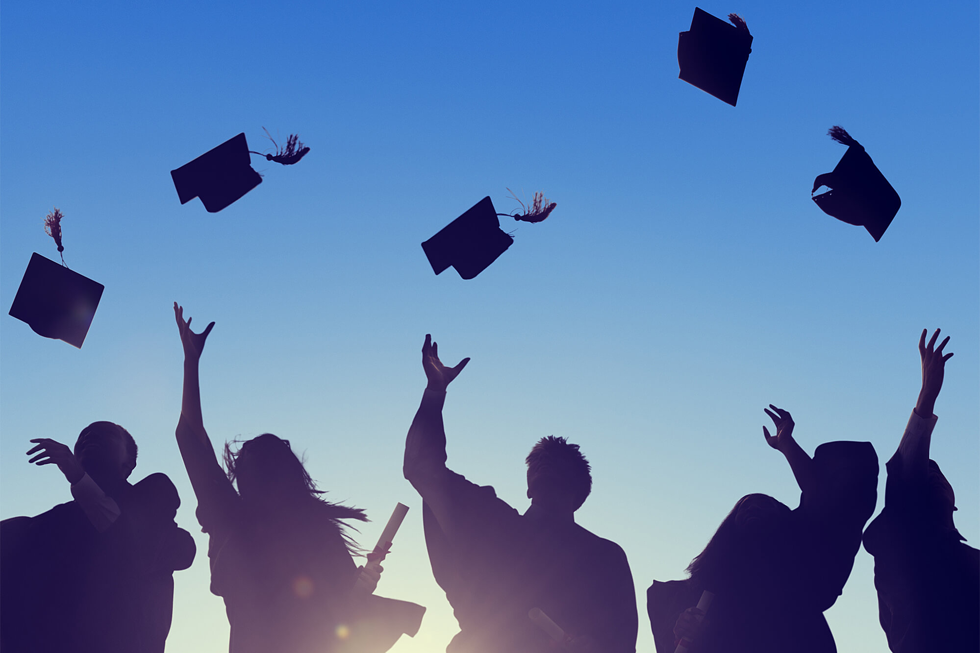 Silouette photograph of graduates throwing their graduation caps up in the air against a blue sky.