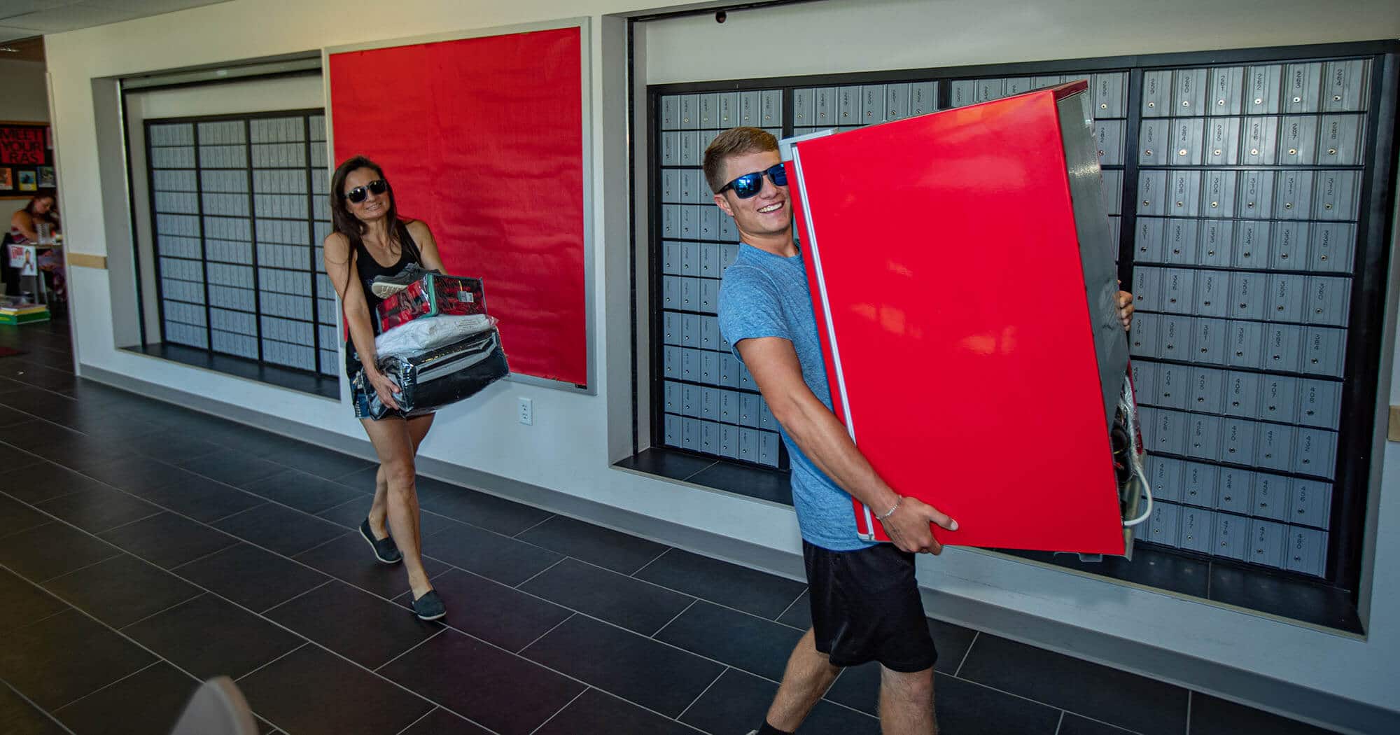 A student carrying a mini-fridge is followed by his mother carrying bedding for his dorm room.