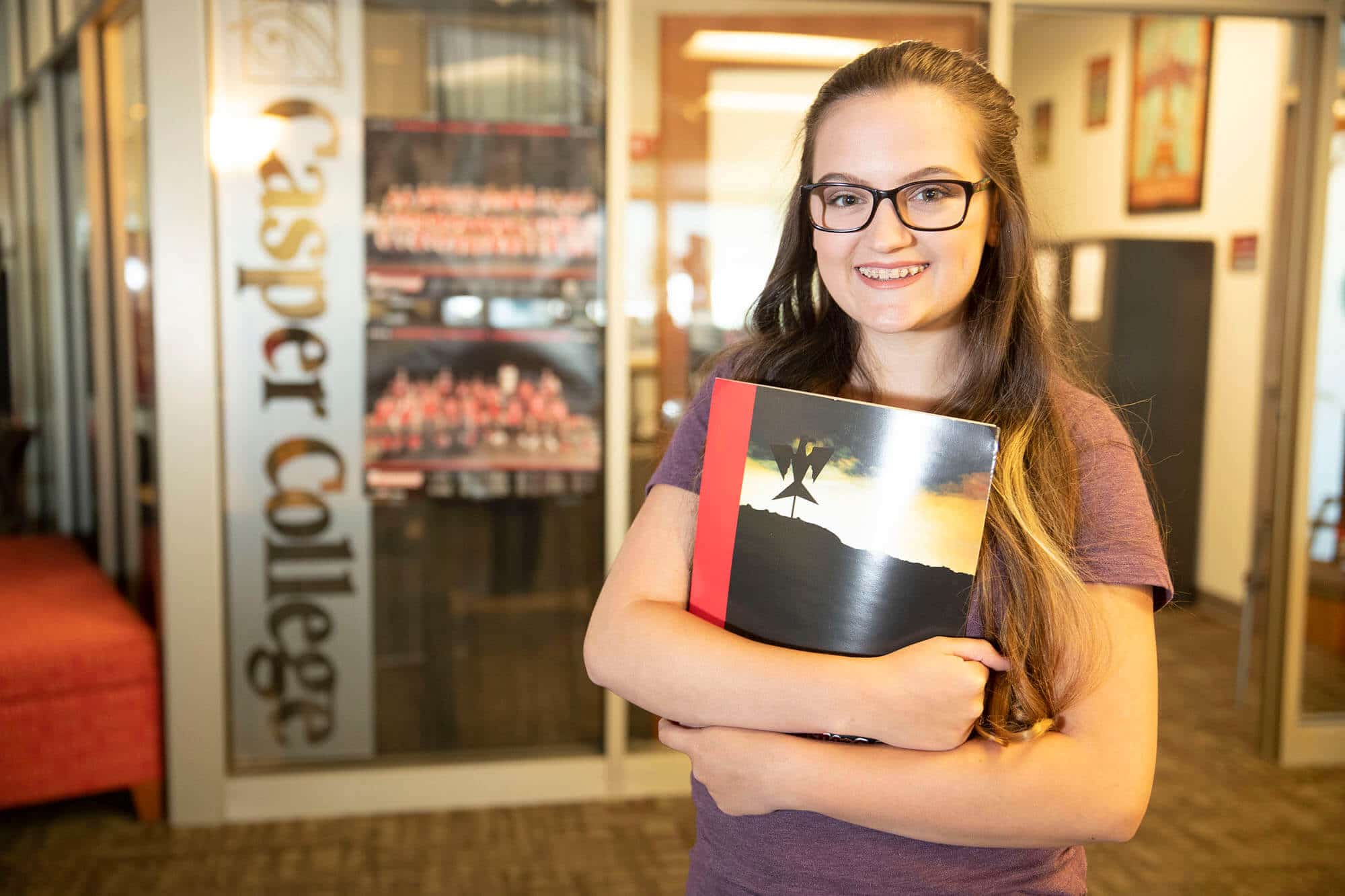 Female student smiling and holding a folder with an office behind her that says Casper College on the window