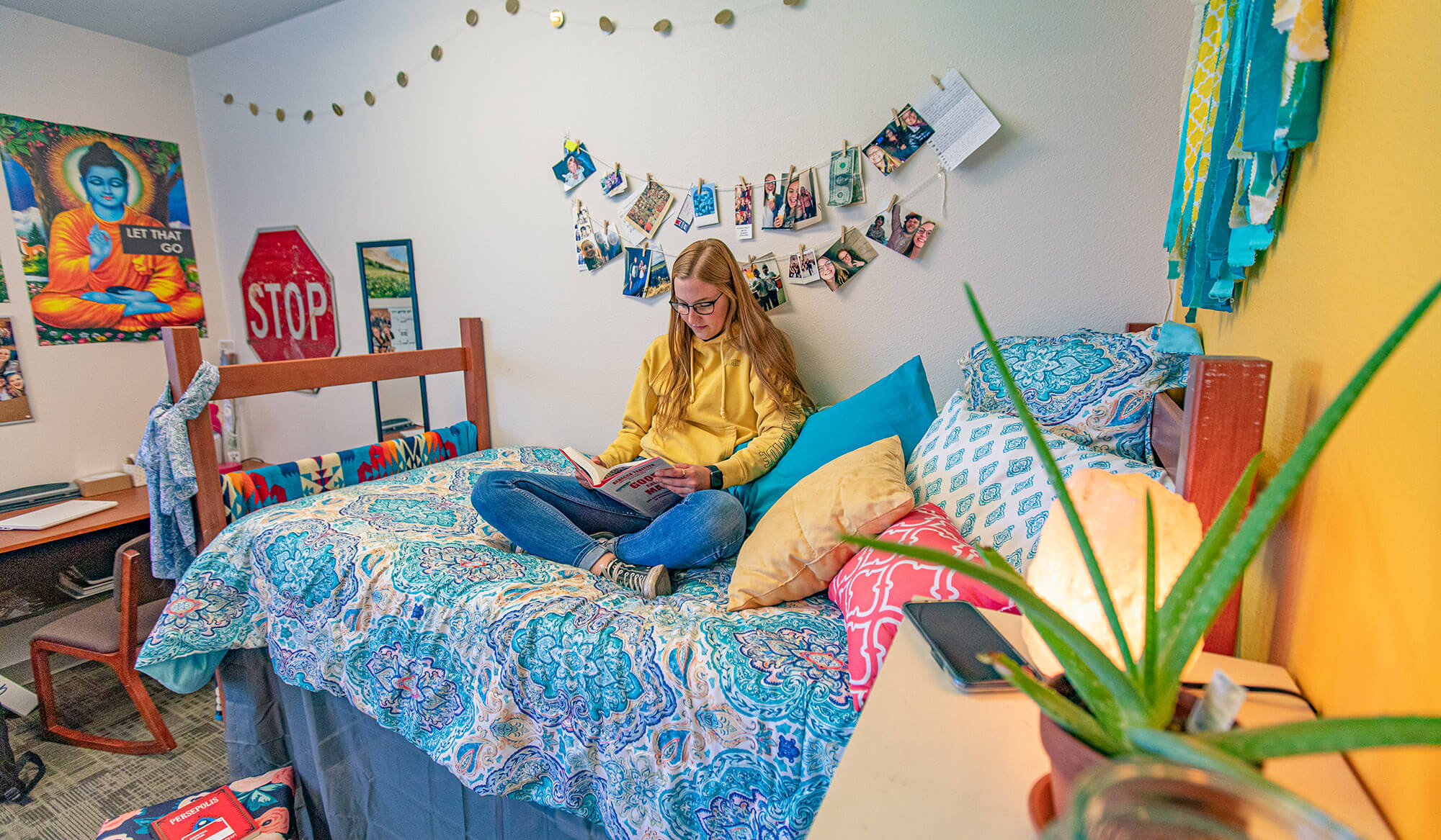 A student reading a book while sitting on the bed of her dorm room; her desk is in the background.