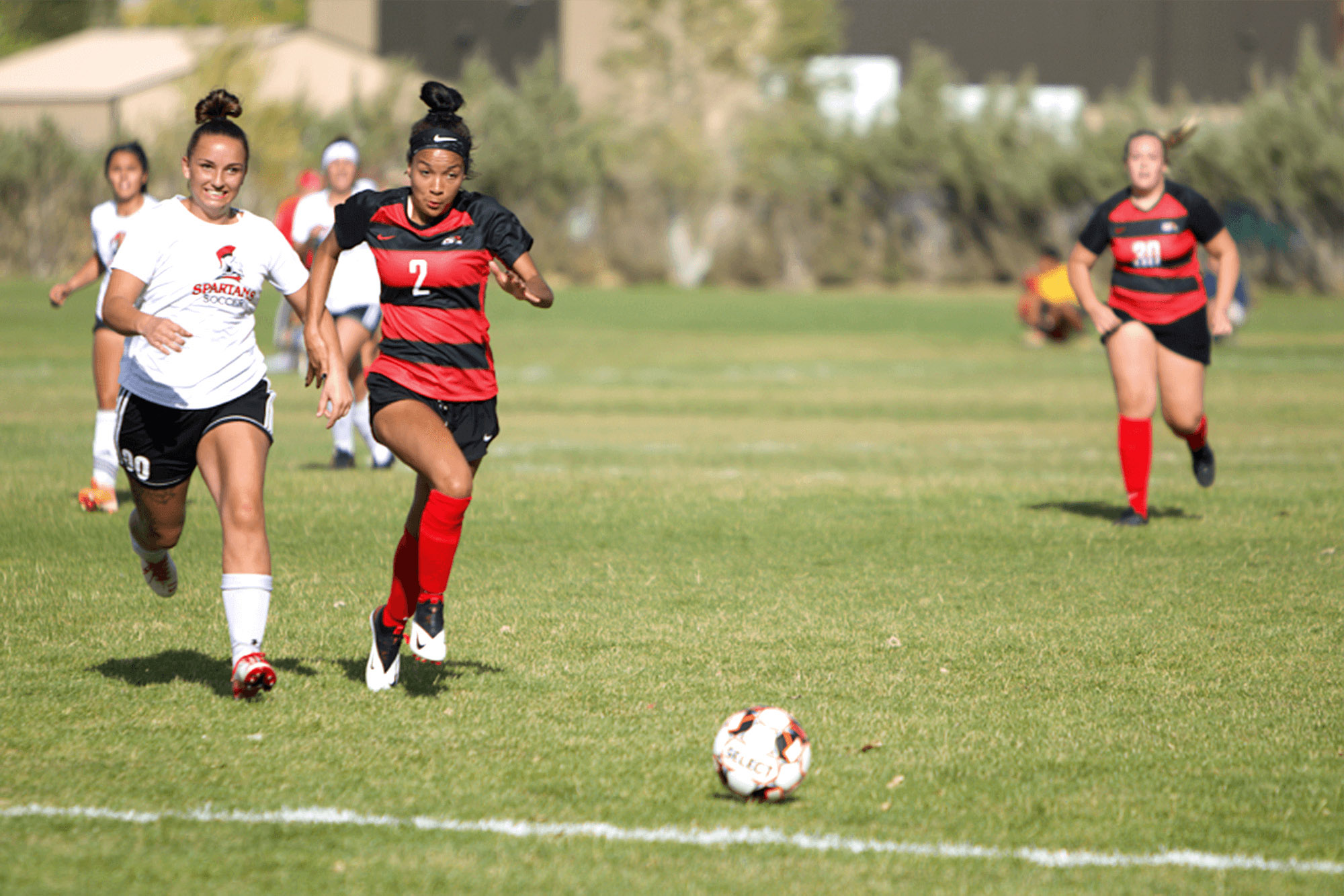 Three female soccer players running after a ball on the field in front of them.