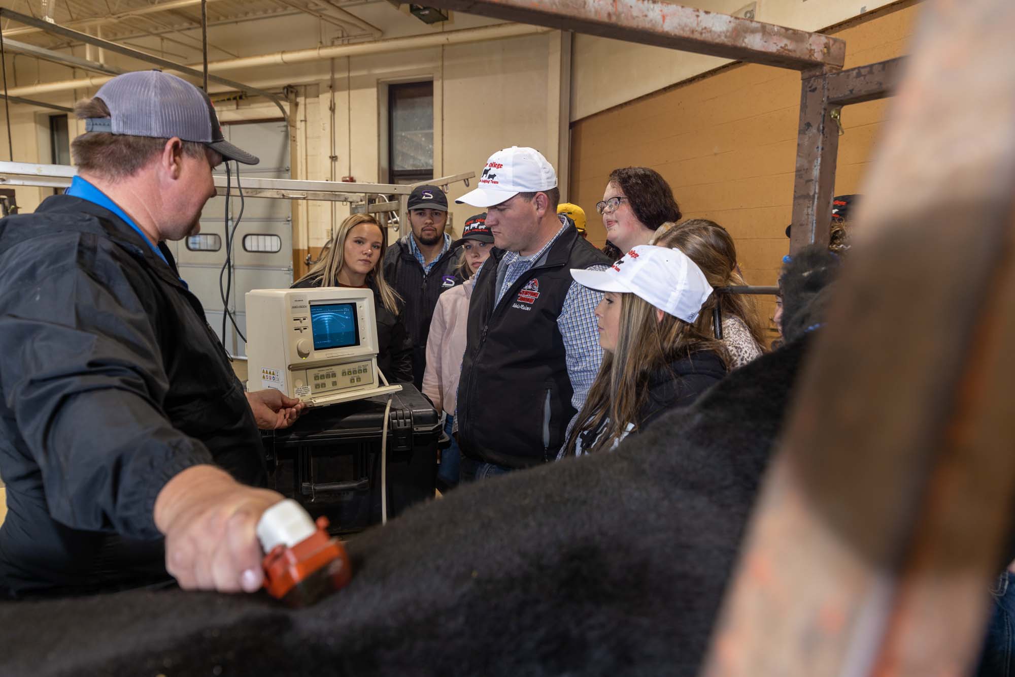 Inside a farm building, a male instructor gestures at a horse while a group of students surround him.