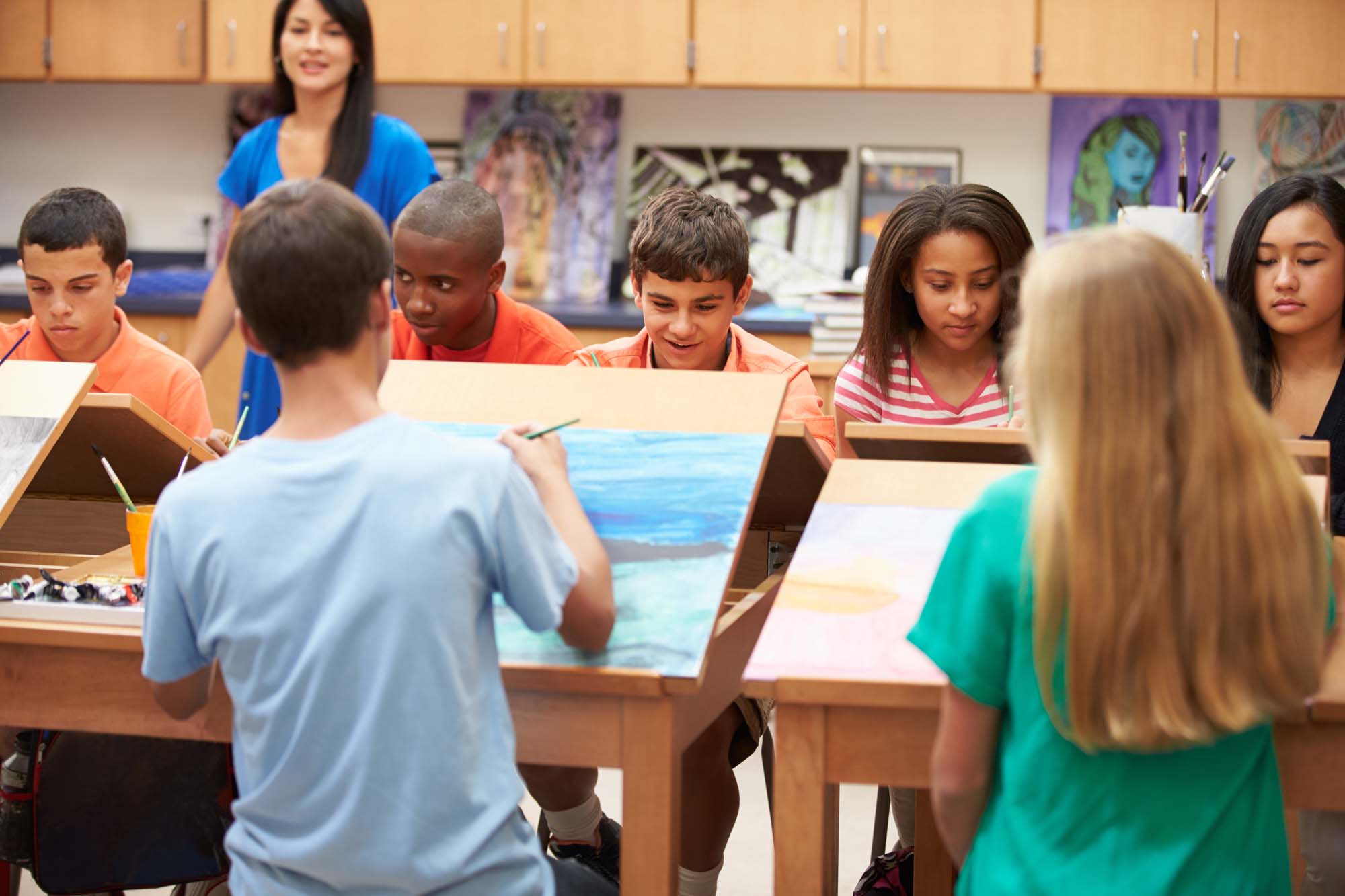 An art instructor provides guidance to a student standing at an easel in a classroom