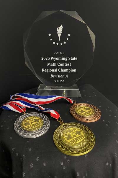 2026-Wyoming-Math-Championship-Awards Photo of a clear octagonal glass trophy engraved ‘2026 Wyoming State Math Contest Regional Champion, Division A’ with a torch and stars logo, displayed behind gold, silver, and bronze medals with red, white, and blue ribbons on a black cloth background.