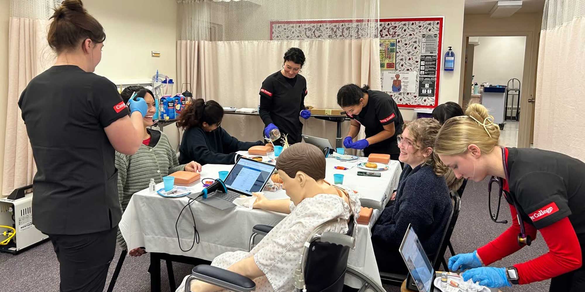 Group of Casper College nursing students in black scrubs practicing clinical skills in a lab, working with medical supplies, laptops, and a patient mannequin around a table.