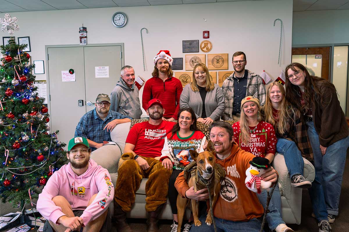 Group photo in a decorated office for the holidays: several people gathered around a couch near a lit Christmas tree, with festive sweaters and a brown dog sitting front and center.