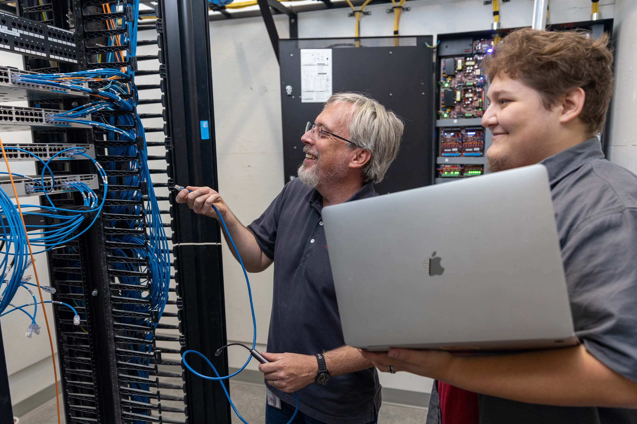 An instructor teaching cybersecurity to a student in front of a server rack.