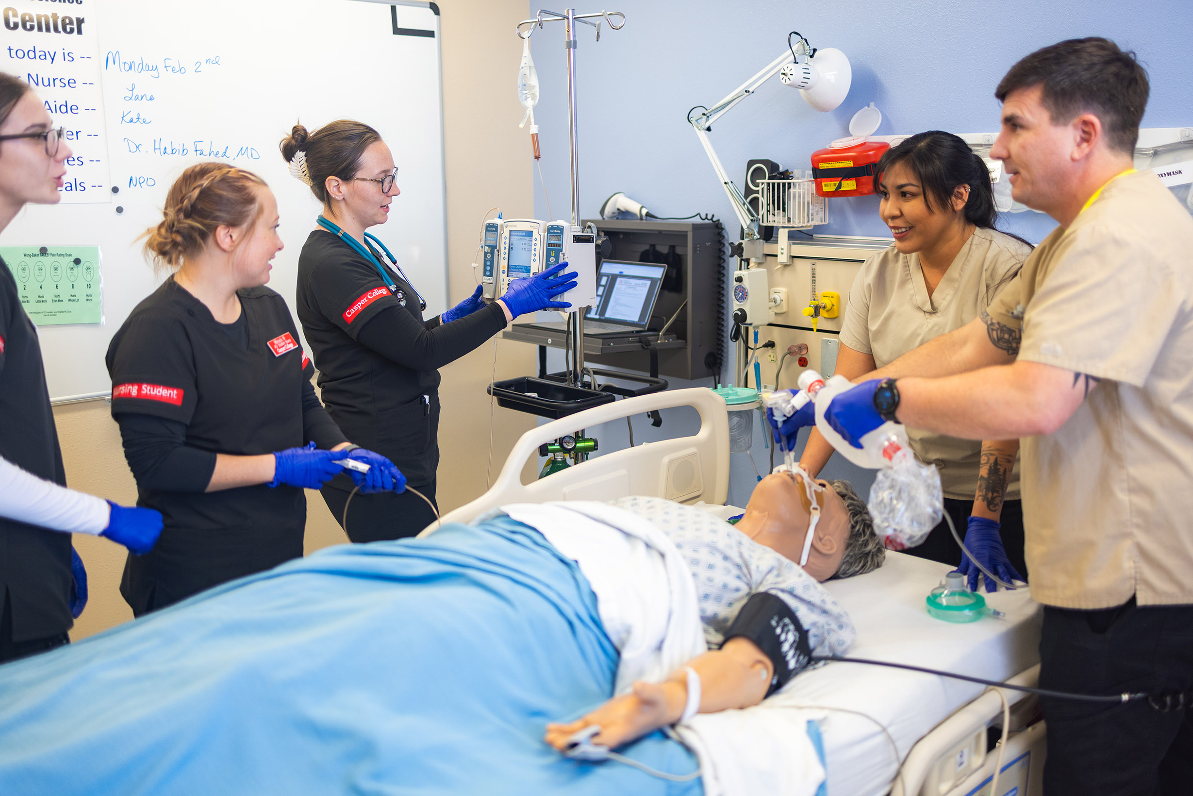 A group of nursing students and an instructor practice a medical simulation in a clinical skills lab. A patient mannequin lies in a hospital bed while one student uses a bag-valve mask to assist breathing. Another student adjusts an IV pump, and others observe and discuss the procedure. The room includes medical equipment, monitors, a laptop, wall-mounted supplies, and a whiteboard with handwritten notes in the background.