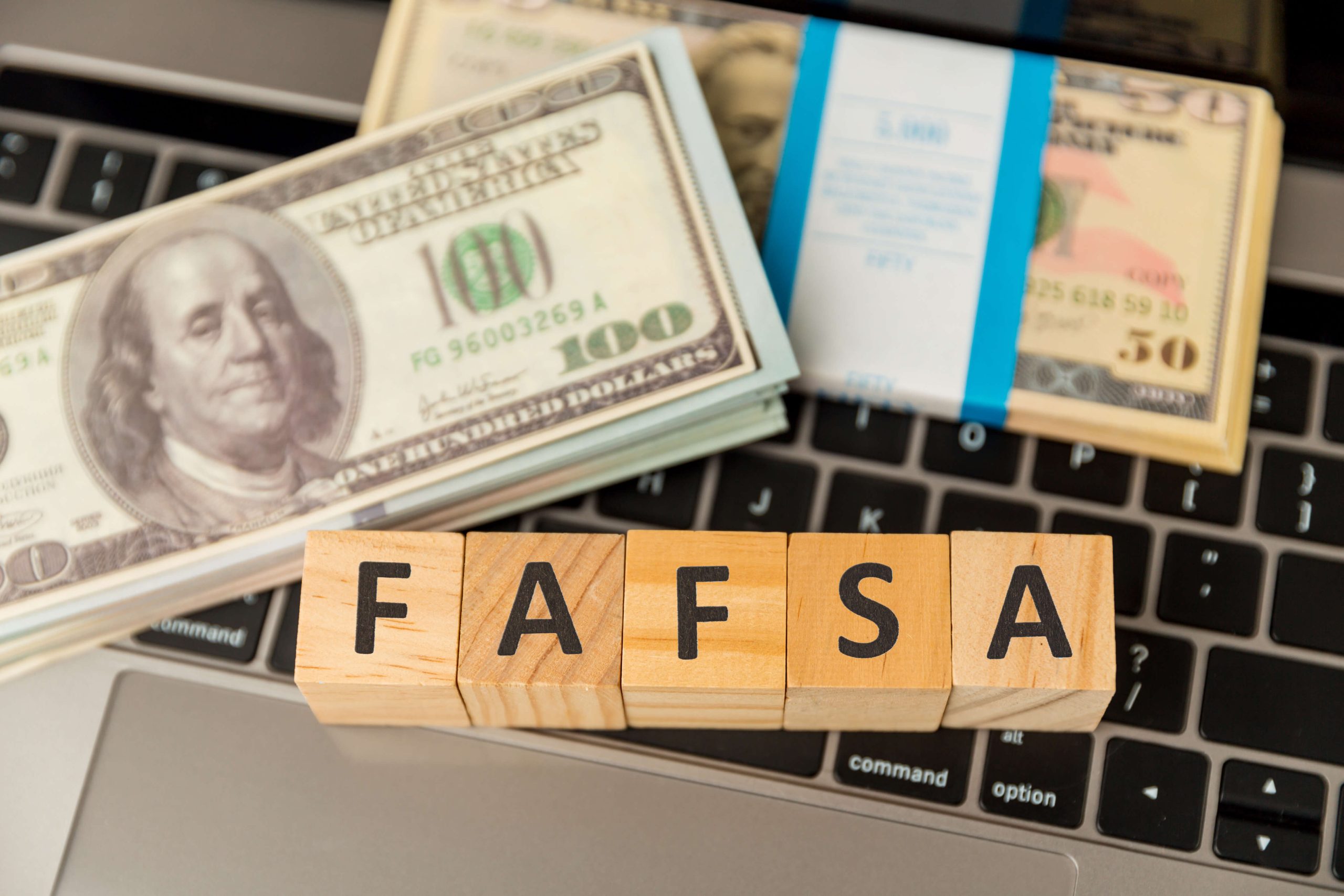 Wooden blocks spelling FAFSA on a laptop keyboard next to stacks of $100 and $50 bills.