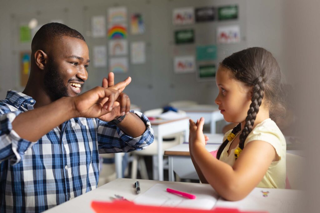 Adult and child smiling while practicing American sign language across a table in a classroom.