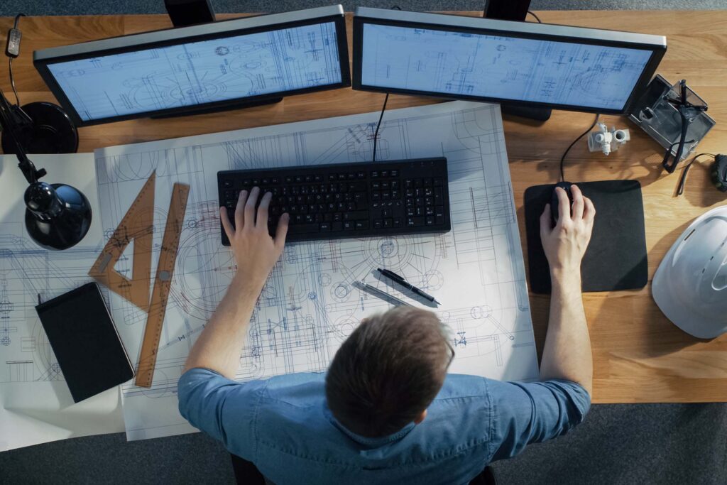 Overhead view of a person working at a desk with monitors, blueprints, and drafting tools in Architectural Graphics and Design.