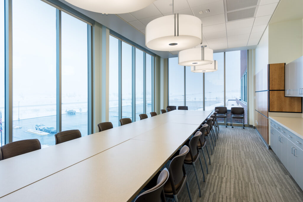 Empty conference room with a long table and chairs, large windows, and overhead lighting, overlooking a snowy outdoor parking area.