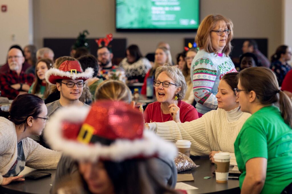 Group of people seated at tables during a festive indoor gathering, talking and smiling while wearing holiday-themed clothing.