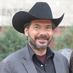 A headshot of a smiling man with a beard and mustache wearing a black cowboy hat, a dark button-down shirt, and a grey blazer.