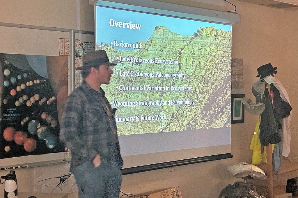 Zach Tennery giving a presentation on Late Cretaceous ecosystems with a projector screen showing Wyoming stratigraphy.