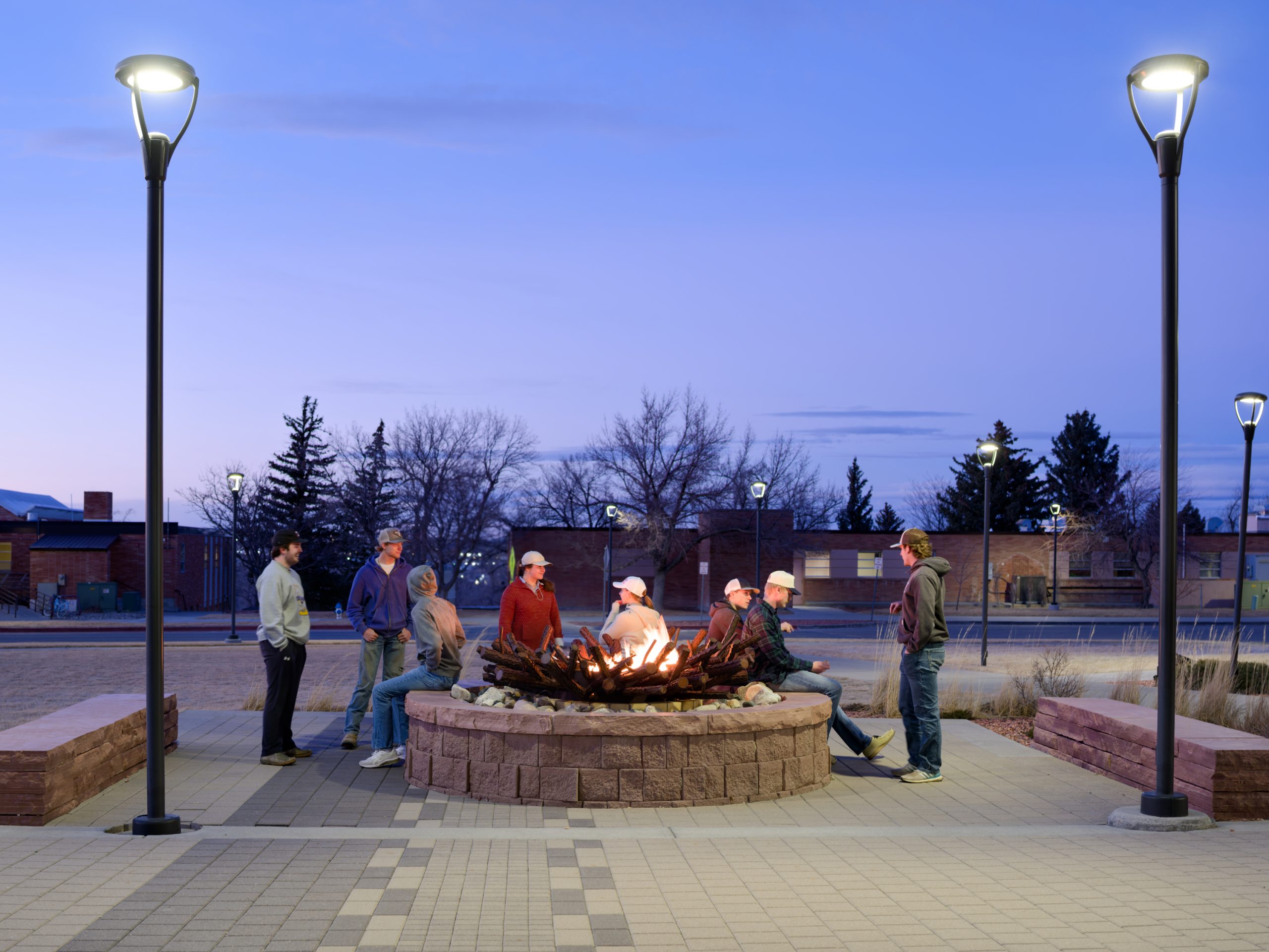 Students standing around the firepit in front of the Union/University building at dusk with the fire lit.