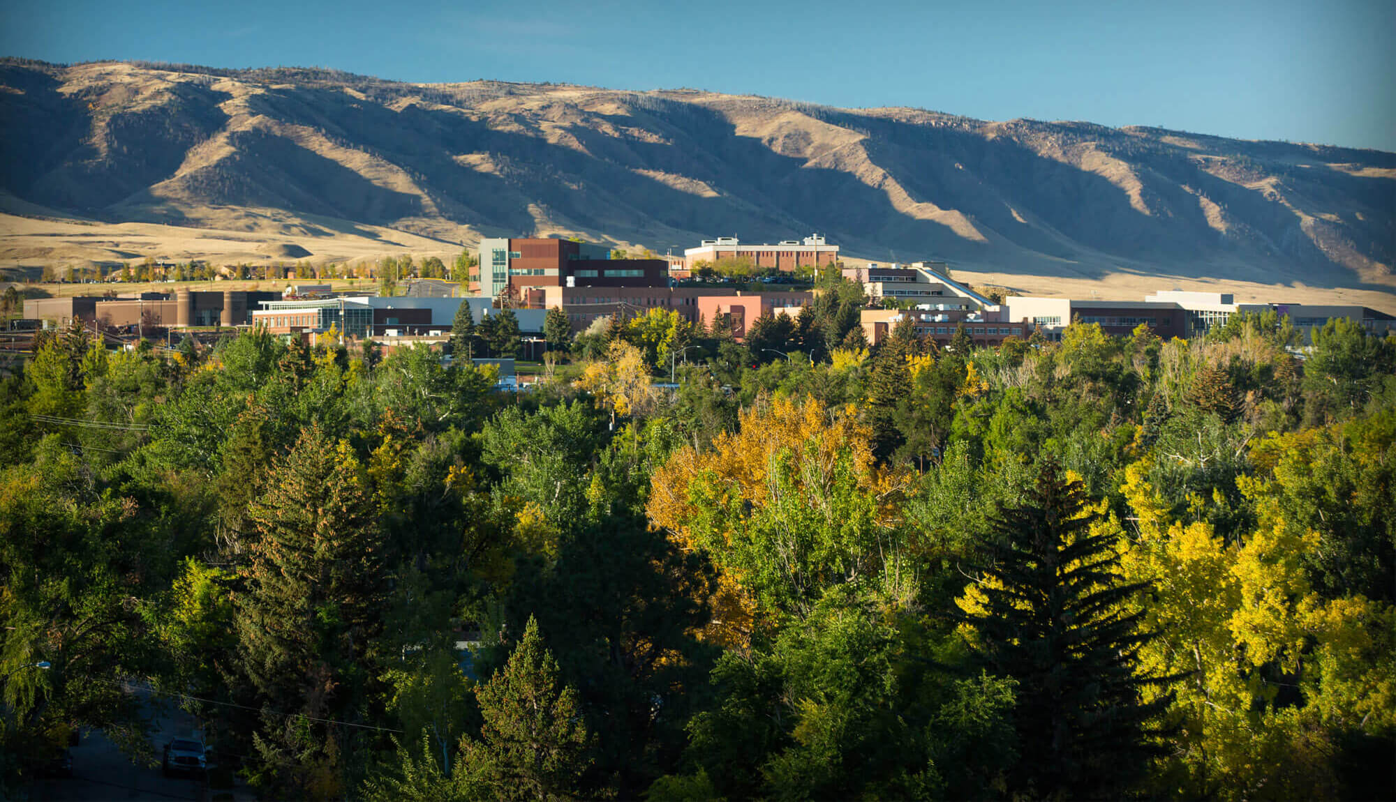 Panoramic view of Casper College buildings nestled against a mountain backdrop with lush green and yellow autumn trees in the foreground.