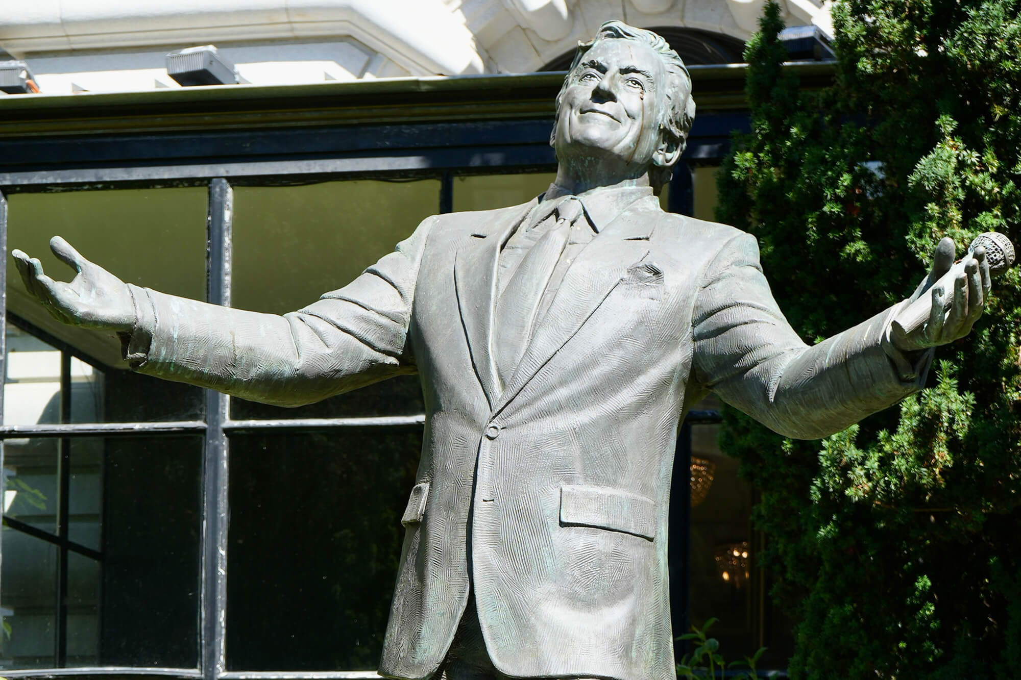 Bronze statue of a smiling male singer with arms outstretched holding a microphone in front of a glass building.
