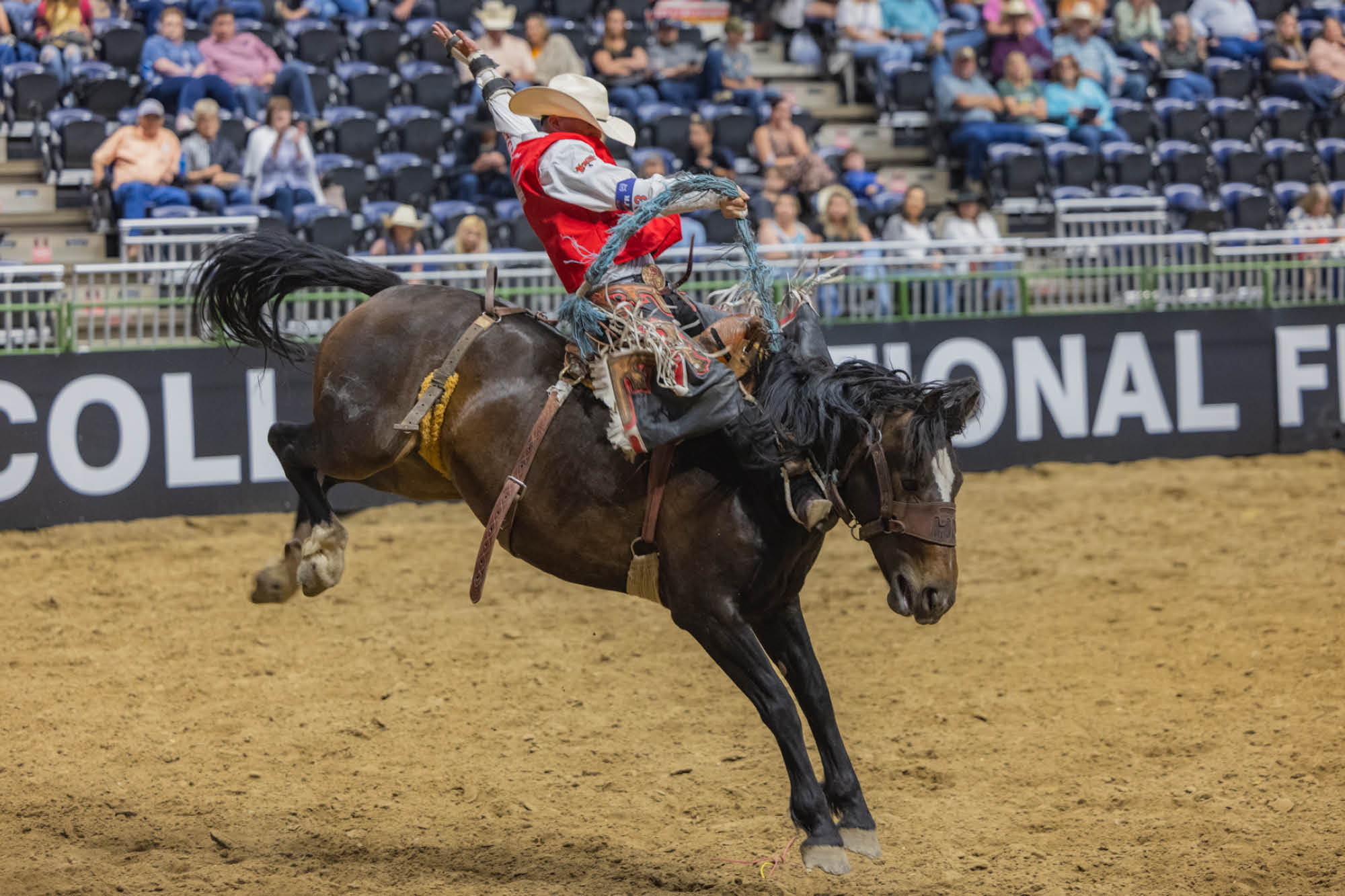A rodeo athlete in a red vest and cowboy hat riding a bucking horse in an arena.