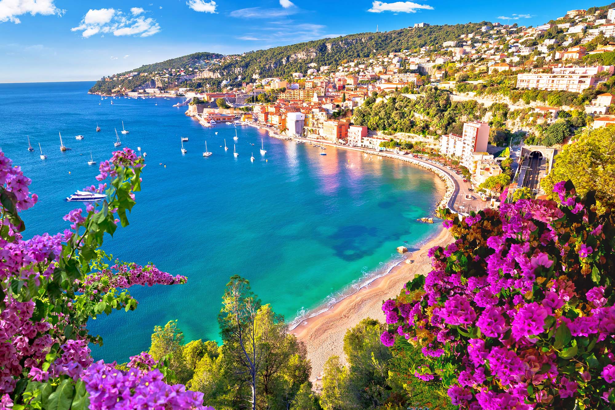 Panoramic view of the French Riviera coastline with blue water, boats, and colorful hillside buildings.