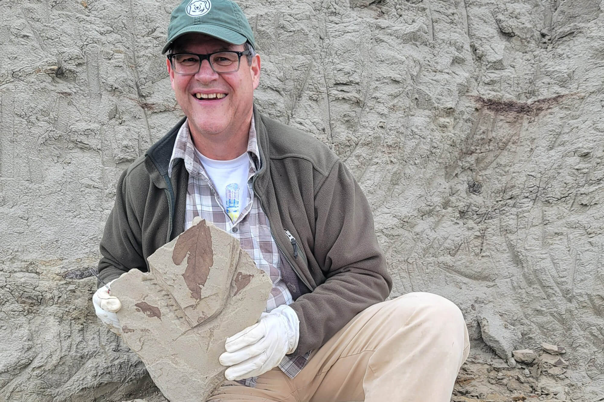 kirk.jphnson.image.0526 Kirk Johnson, Ph.D., kneeling outside and holding a stone slab containing a clear prehistoric leaf fossil specimen.