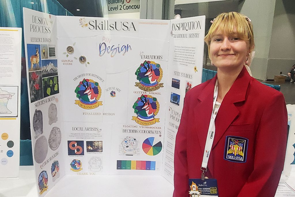 Fionna Kerkes standing next to her tri-fold presentation board detailing her award-winning antelope pin design.