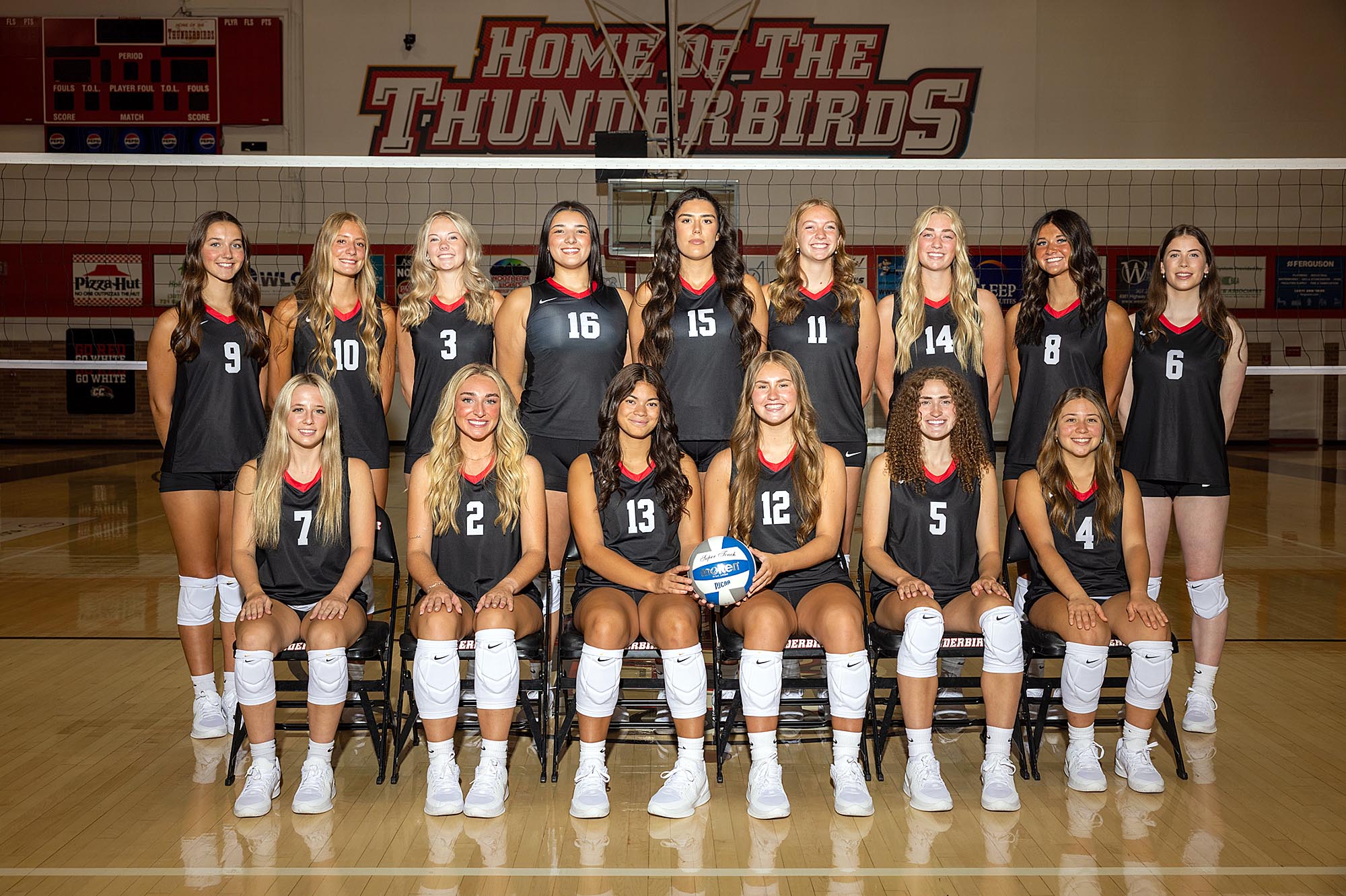 Casper College women's volleyball team posing together on their home court under a "Home of the Thunderbirds" sign.