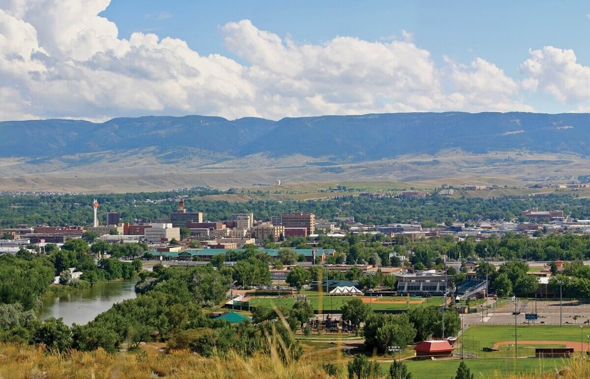 Aerial view of a city in the foreground with lots of green trees and a mountain in the distance with blue skies and white puffy clouds.