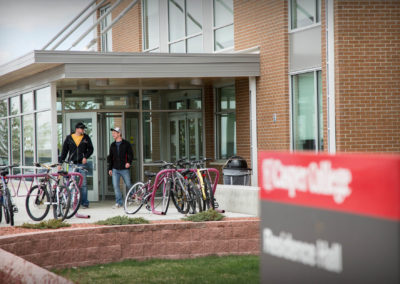 Two students walk out of the entrance of the Residence Hall. Nearby, two bike racks are filled with bikes.