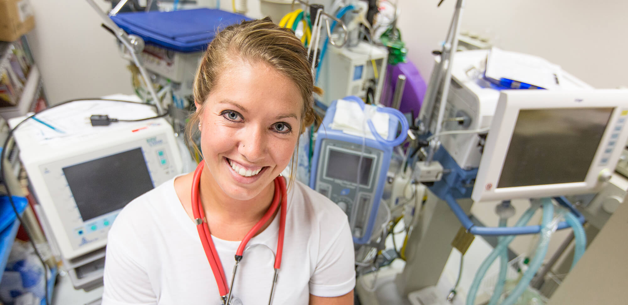 photo of a student wearing scrubs with a stethoscophe around neck and medical equipment in background