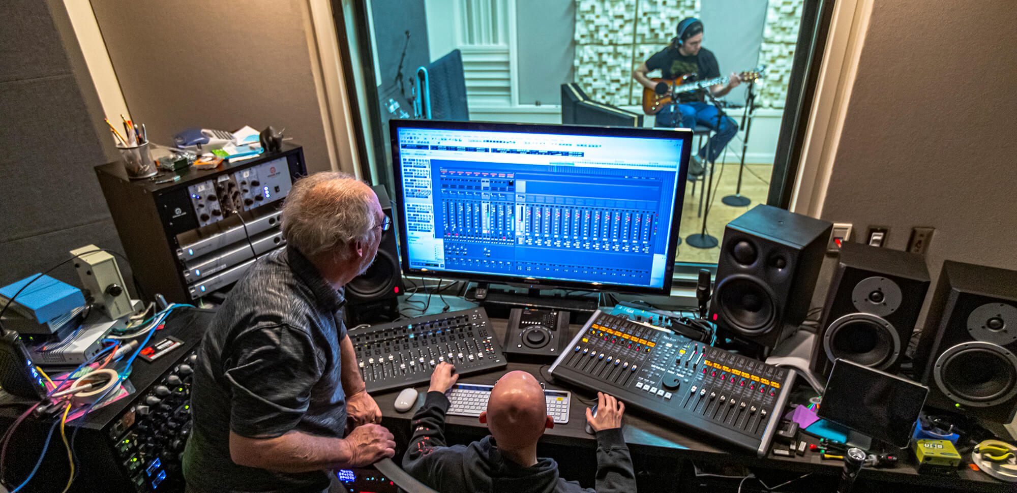 An instructor oversees a student working at a mixing board while anther student plays the guitar in a practice room.