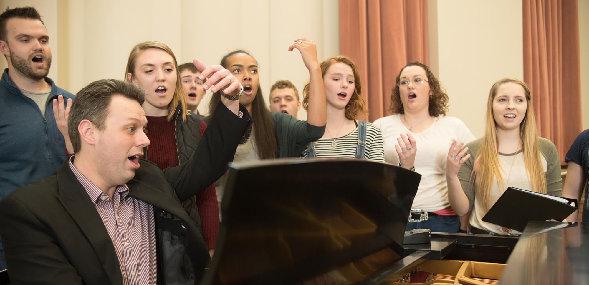 Students practice vocal exercises while gather around an instructor playing the piano.